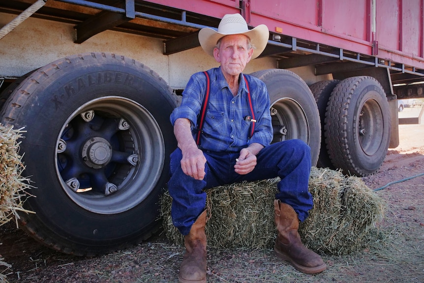 An older man wearing a cowboy hat and boots sits on a hay bale in the shade of a truck.