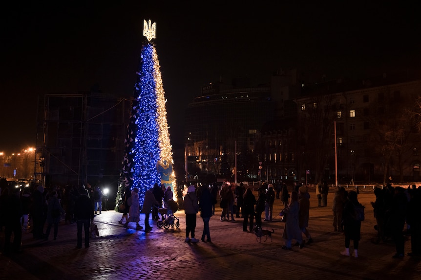 People gather around Christmas tree decorated in blue and yellow lights in a town square in Kyiv.
