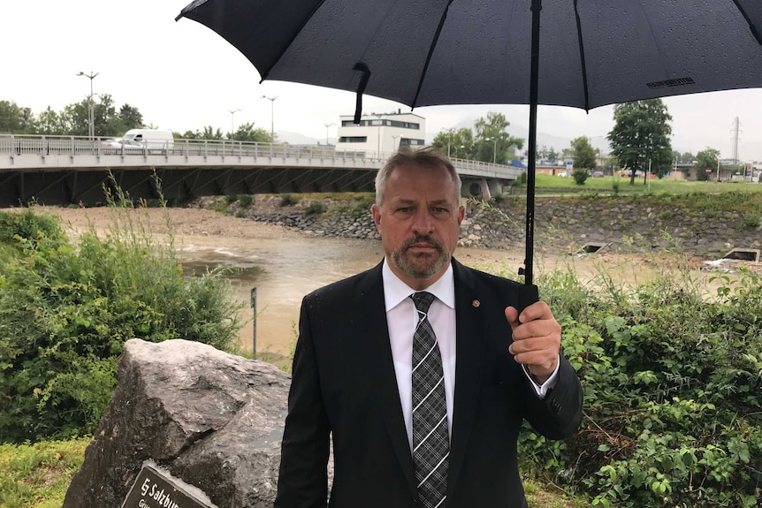 A man wearing a black suit, white shirt and tie holds an umbrella while standing in front of a bridge over a river