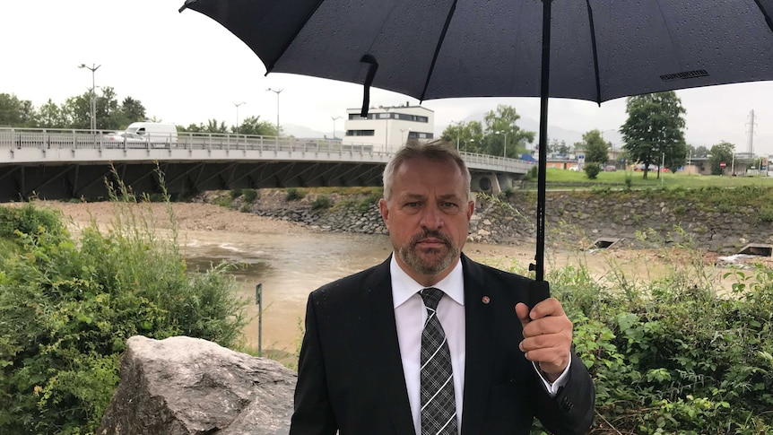 A man wearing a black suit, white shirt and tie holds an umbrella while standing in front of a bridge over a river