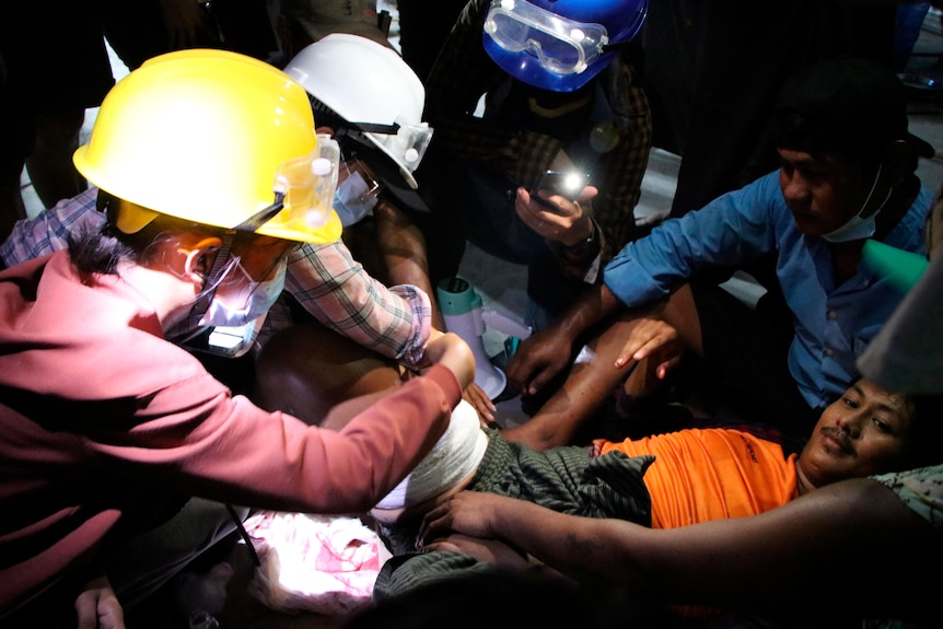 Anti-coup protesters wearing helmets attend to a man with a bandage on his right leg.