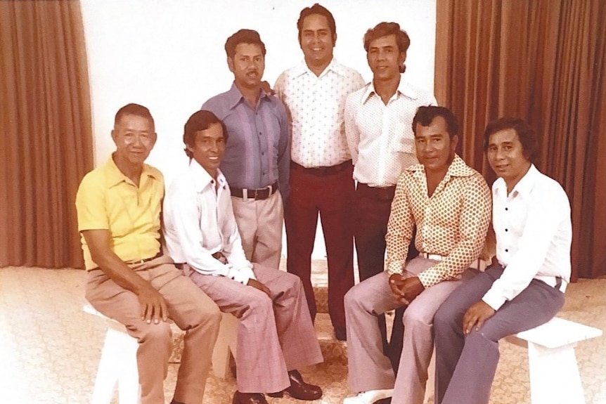An old colour photo of seven men standing in a semi circle, some sitting and some standing