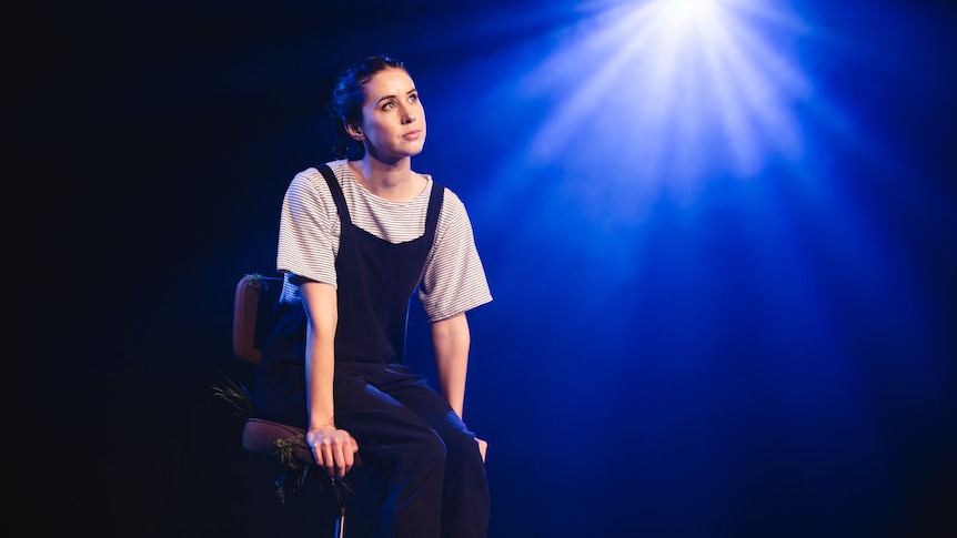 A brunette woman in her 30s, wearing overalls, stares off into the middle distance, while sitting on a chair on stage. 