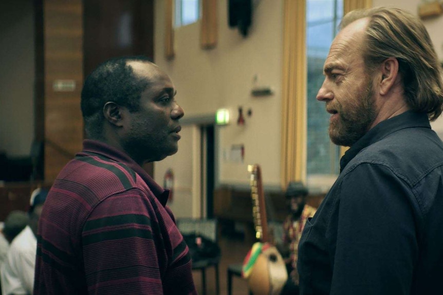 Actors Andrew Luri and Hugo Weaving in the film Hearts and Bones, looking at each other tensely in a community hall