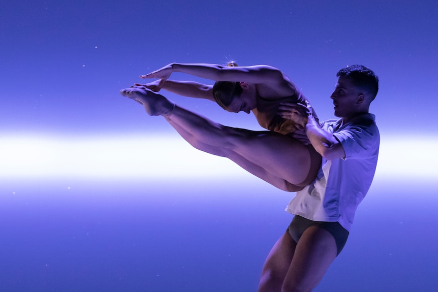 A male dancer lifts a female dancer, who bends to touch her toes. There is bright blue light behind them.
