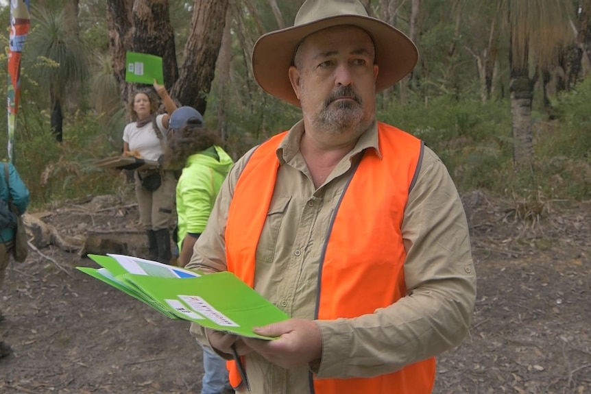 Man standing in forest holding green folder wearing high-vis vest and wide brimmed hat