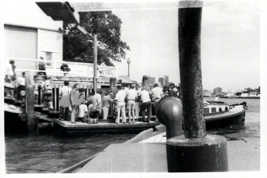A black and white image of a timber vessel on a harbour with a crowd of men standing on board.