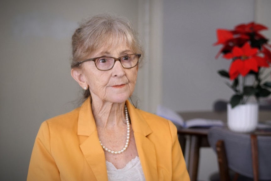 A woman with glasses in a yellow jacket. Table with red flowers in background