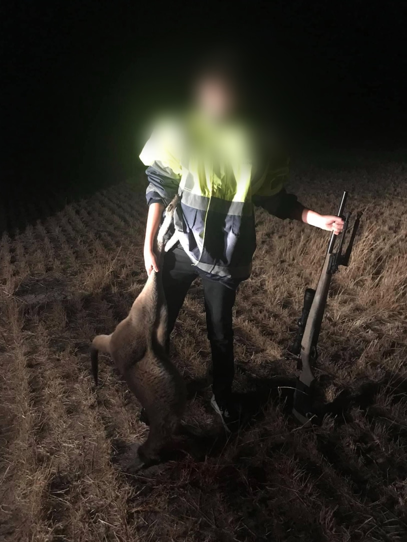A child holds a gun and kangaroo carcass as he stands outside in the dark, light by a spotlight.