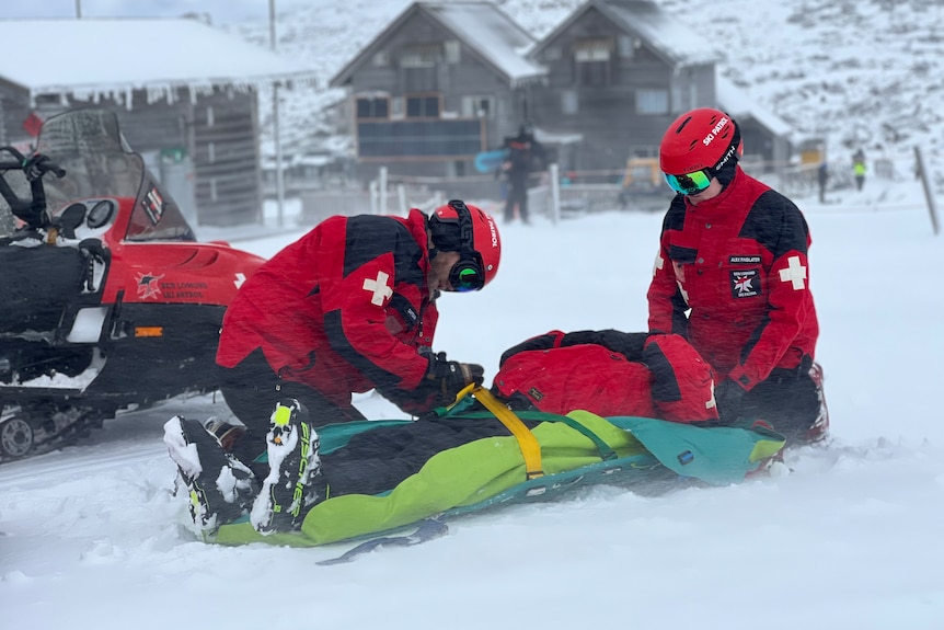 Two people wearing red snow jackets administer to another lying on a stretcher in the snow.