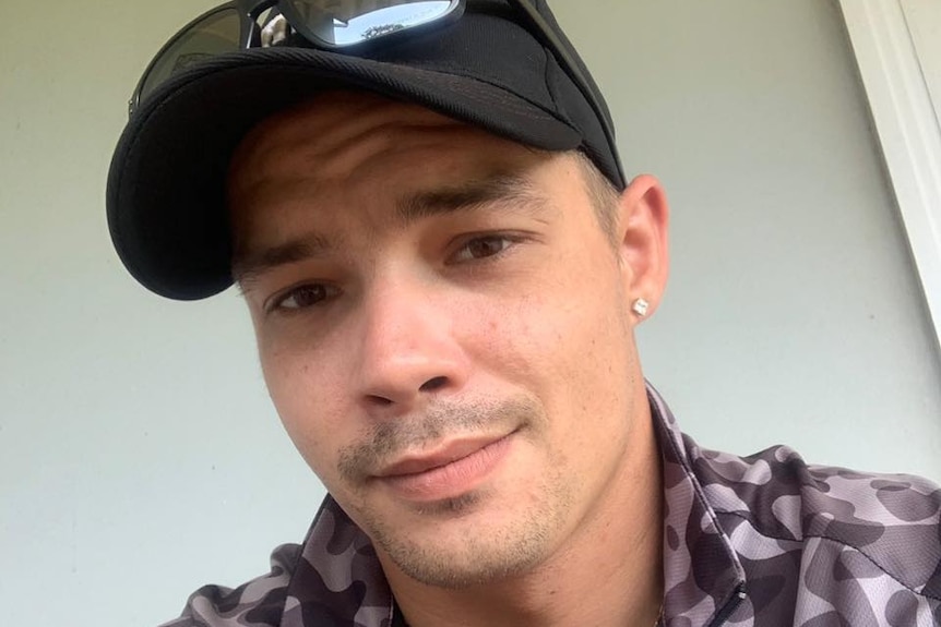 A selfie image of a man wearing a black cap, a chequered shirt and with a pierced ear