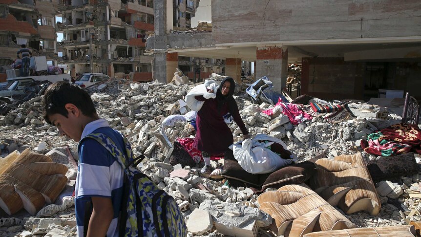 An earthquake survivor carries her belongings over debris in front of her house.
