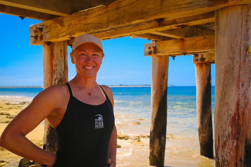 a woman wearing bathers standing underneath a pier on a beach.