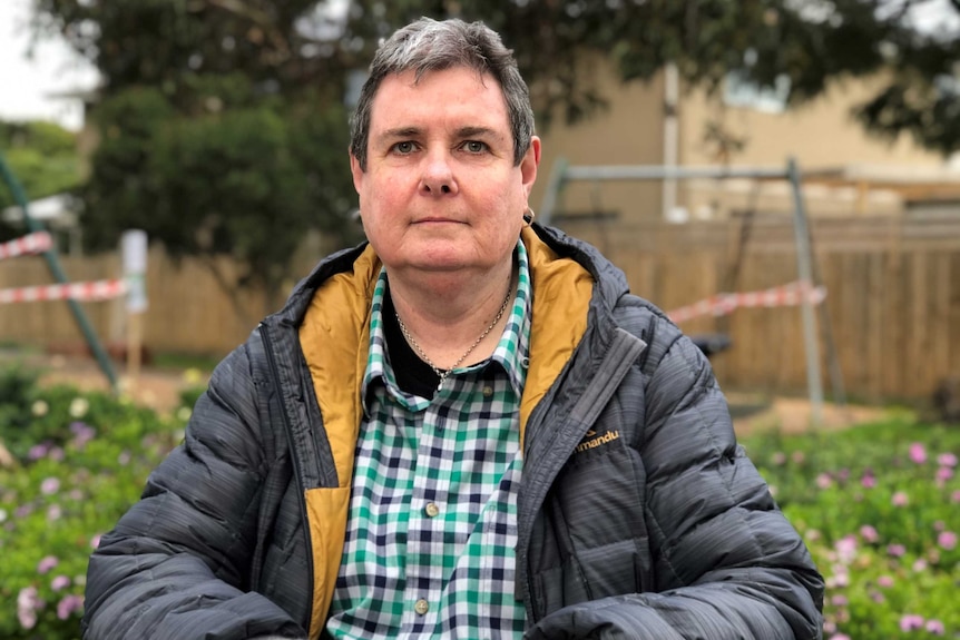 A portrait of Julie Dempsey wearing warm clothing and a puffer jacket, sitting in a park with pink flowers in the background.