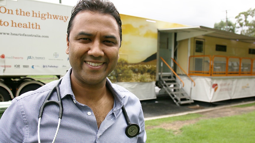A man with stethoscope around his neck smiles, bus with stairs and orange barriers behind him.