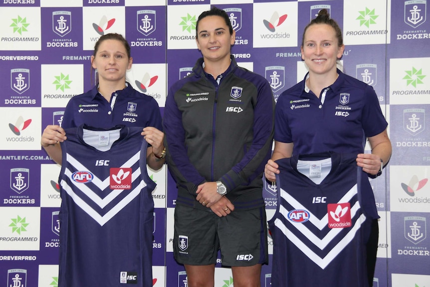 Fremantle Dockers women's players Kiara Bowers and Kara Donnellan stand either side of coach Michelle Cowan holding team jumpers