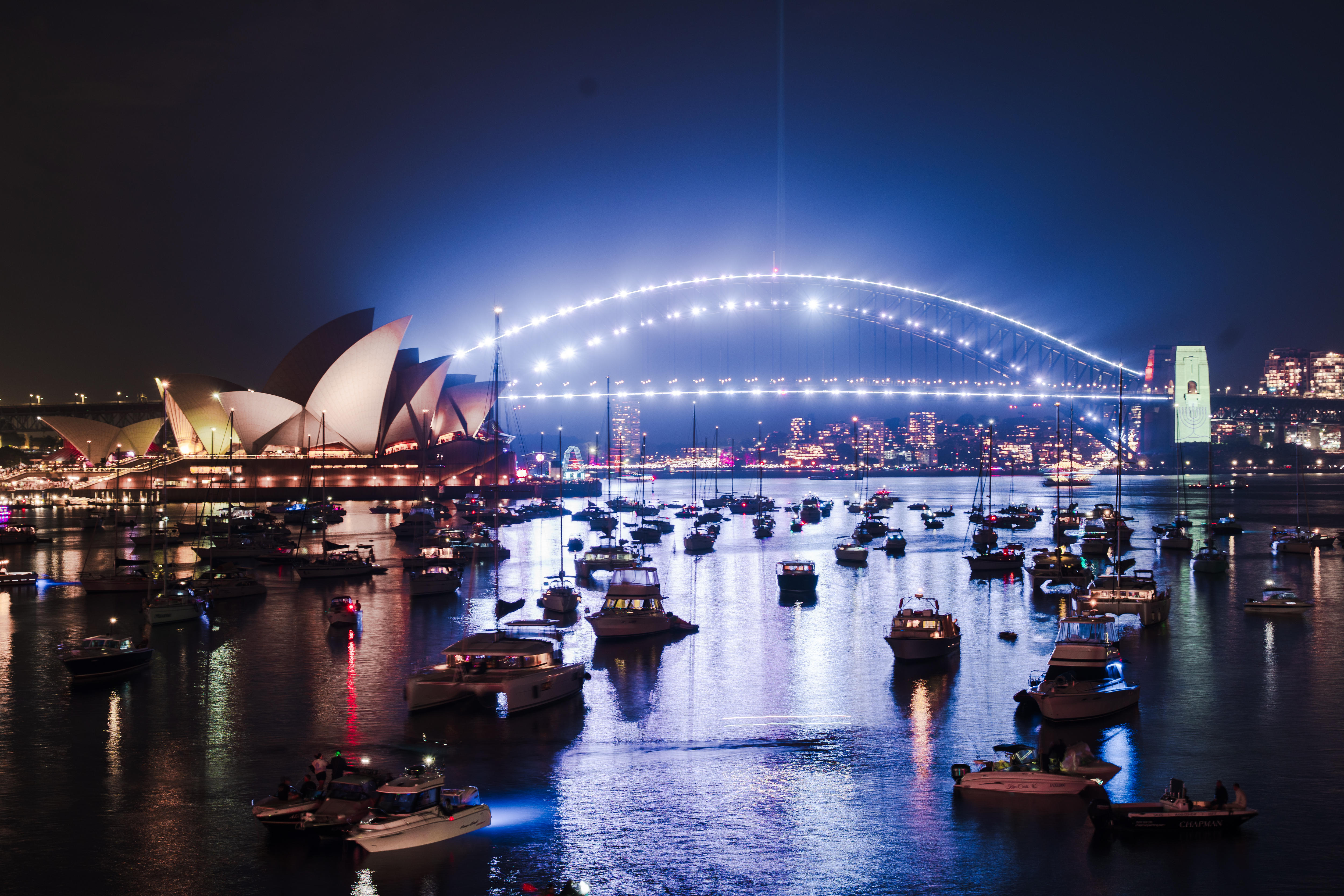 El Puente del Puerto de Sydney iluminado con luces blancas con una menorá en los pilones y barcos en el agua.