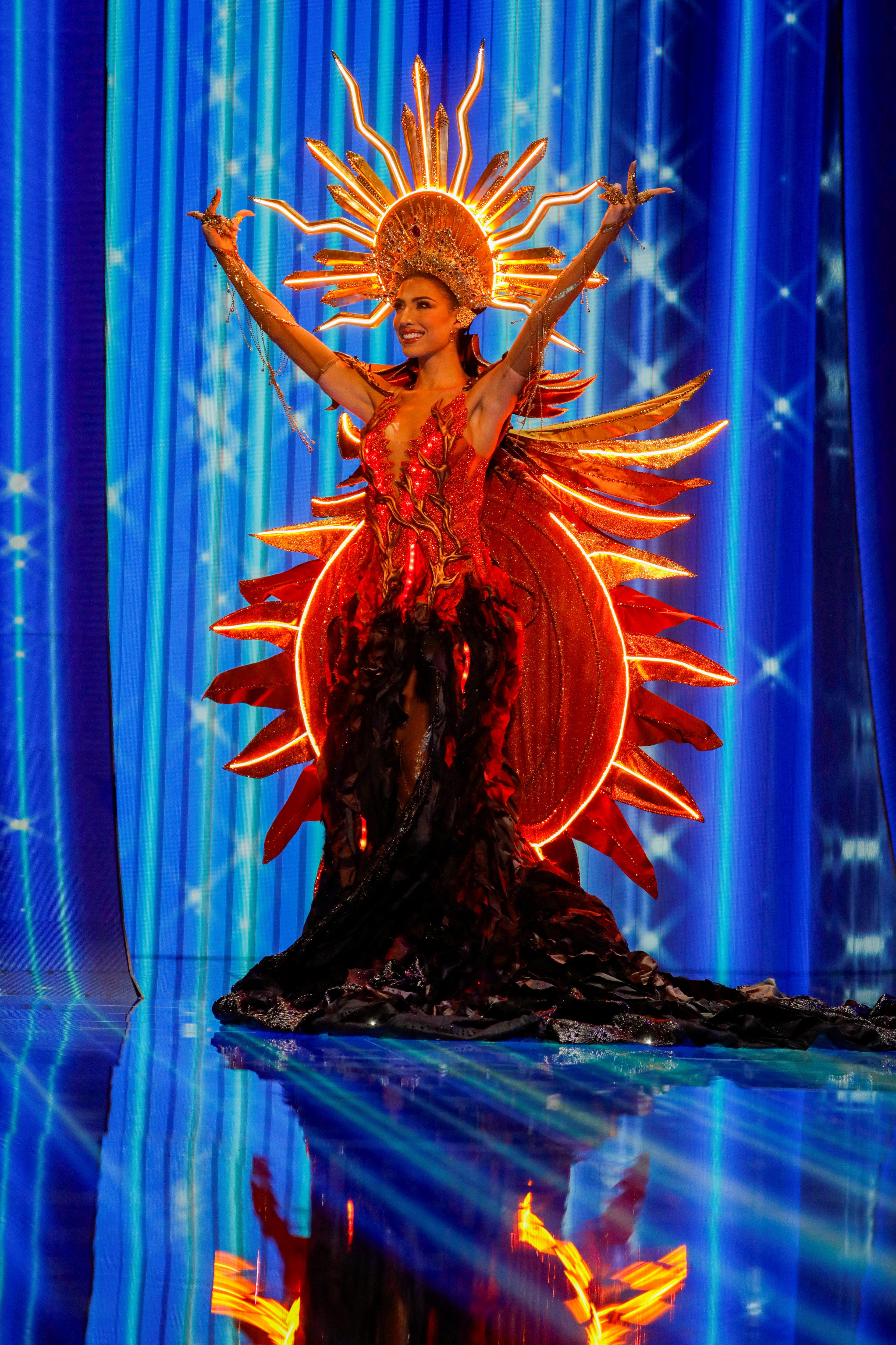 Isabella Garcia-Manzo wearing a headdress that looks like the sun and a long dark red gown, all illuminated