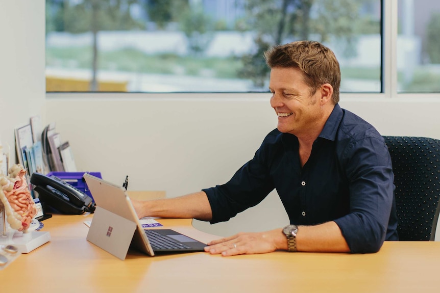 Man sits at desk in front of large window, looking at laptop screen and smiling