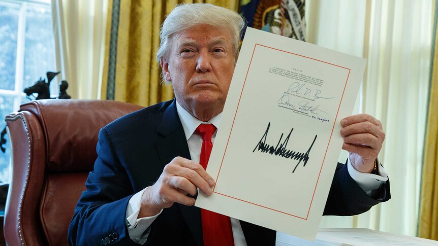 Donald Trump holds tax bill while sitting at his desk in the Oval Office with a serious look on his face