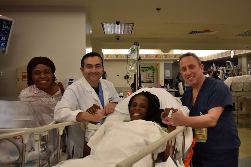 A woman in a hospital bed holding the hand of medical staff, all with big smiles on their faces