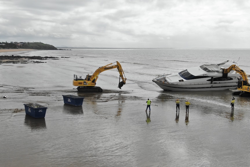 A crane pulls apart a yacht grounded on a beach with skip bins in front of it