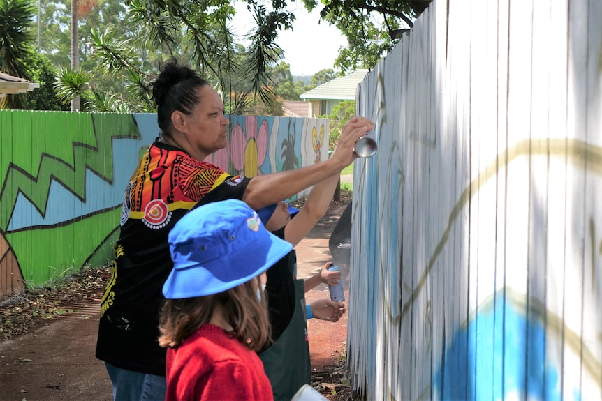 Female artist and children spraypainting mural on fence in lanweay.