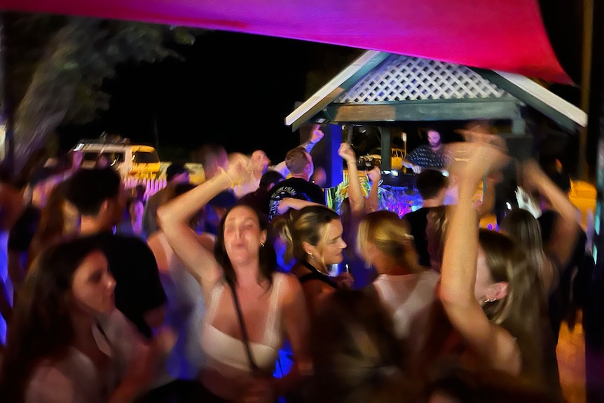 Women dance enthusiastically at an outdoor bar with a DJ visible onstage.