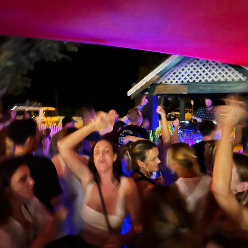 Women dance enthusiastically at an outdoor bar with a DJ visible onstage.