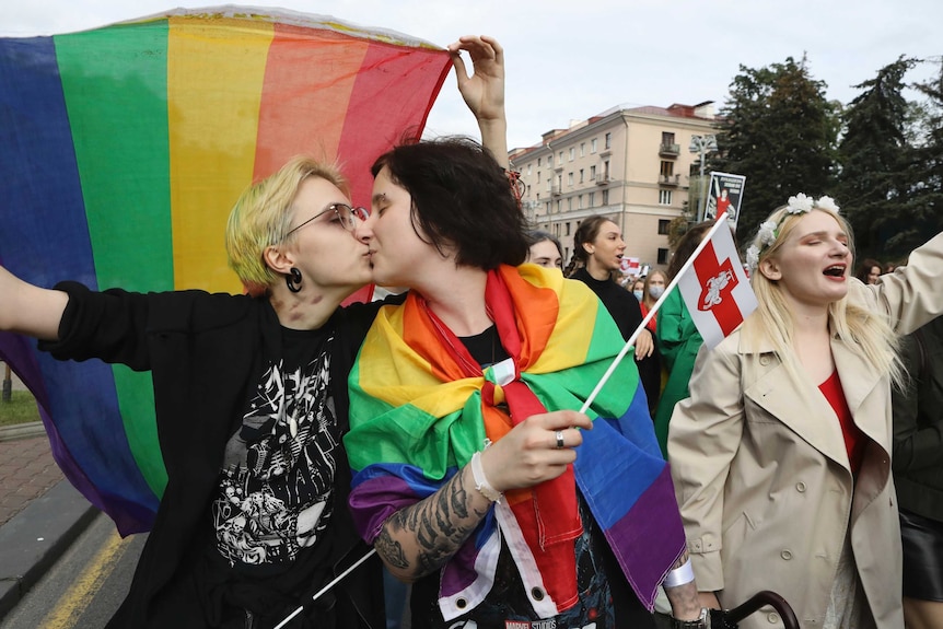 Two young women kiss while one holds a rainbow flag over their heads as the march with a large group of women down a large road.