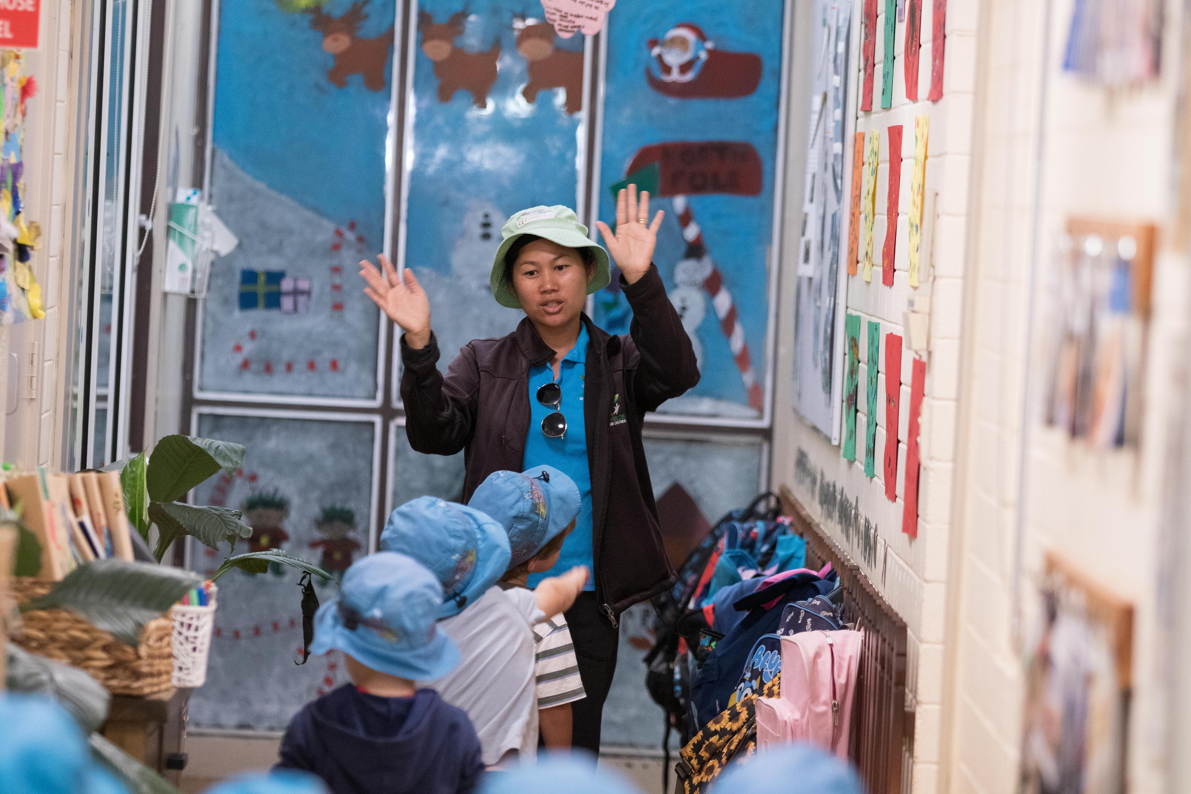 A child care worker waving her hands to get the attention of young children walking down a hallway. 