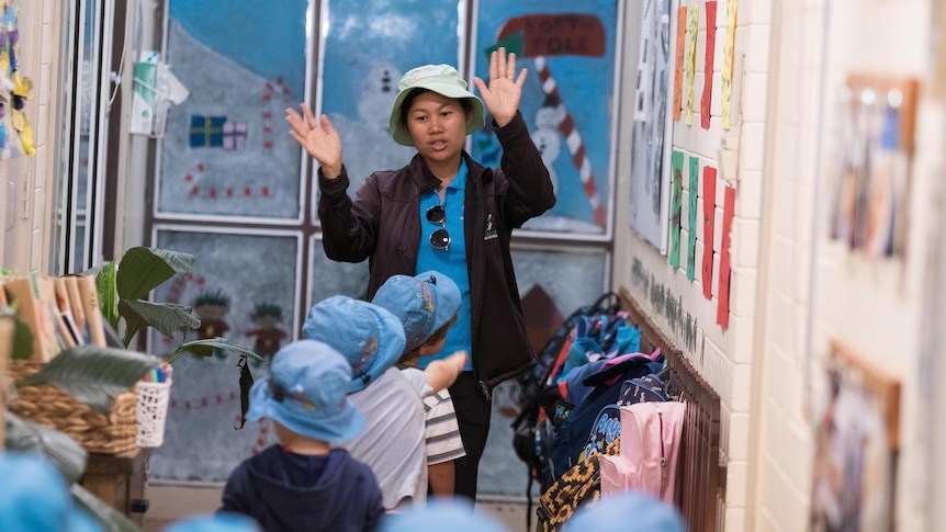 A child care worker waving her hands to get the attention of young children walking down a hallway. 
