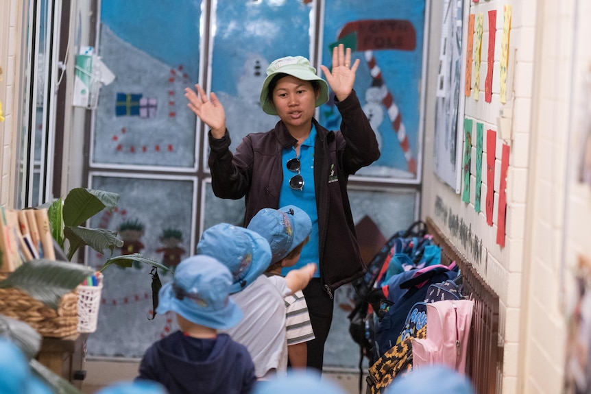 A child care worker waving her hands to get the attention of young children walking down a hallway. 