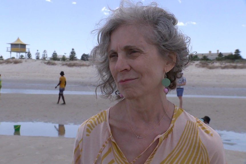 A woman with grey wavy hair wearing a yellow top stands on a beach. Two people walk on the sand behind her