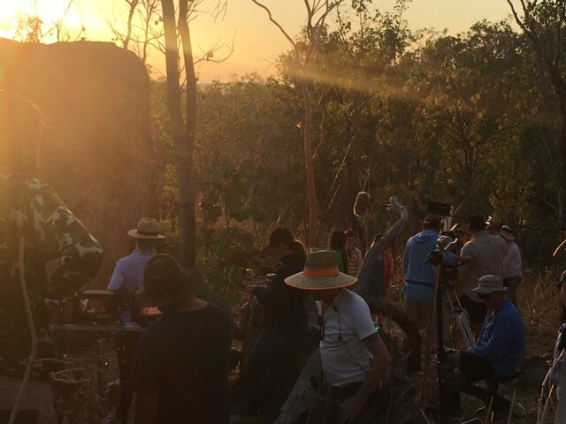 A film set has been set up in bushland overlooking an escarpment. The sun has set and the sky above is orange.