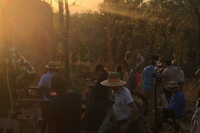 A film set has been set up in bushland overlooking an escarpment. The sun has set and the sky above is orange.