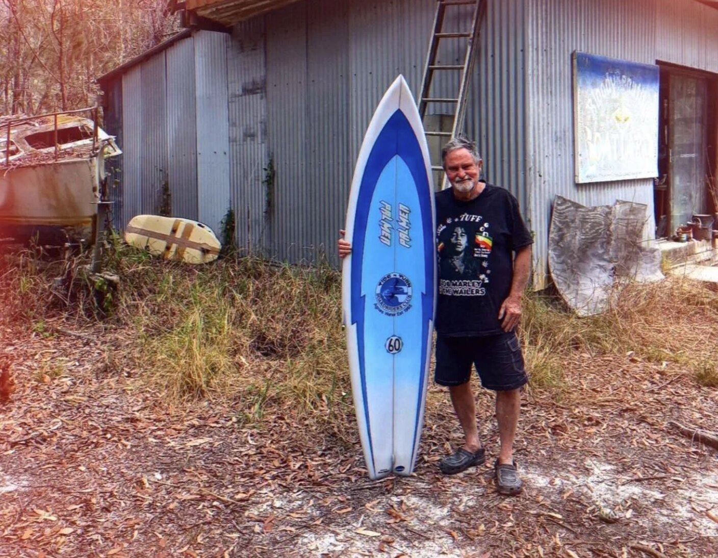 Hombre varado junto a una tabla de surf azul y blanca en posición vertical frente a un cobertizo de hojalata.