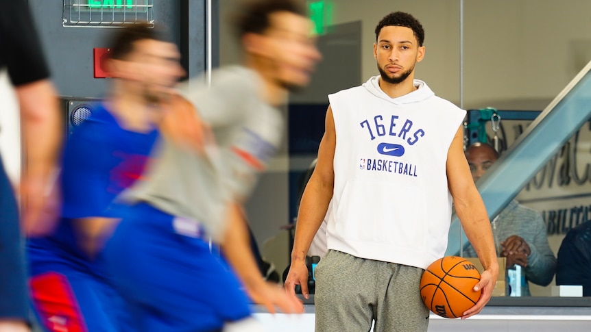 NBA players are a blur of movement at training as a man stands to one side wearing a '76ers basketball' top and holding a ball.