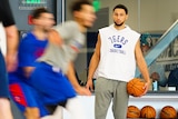 NBA players are a blur of movement at training as a man stands to one side wearing a '76ers basketball' top and holding a ball.