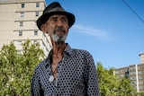 A man wearing a blue shirt and a round hat stands outside large apartment towers