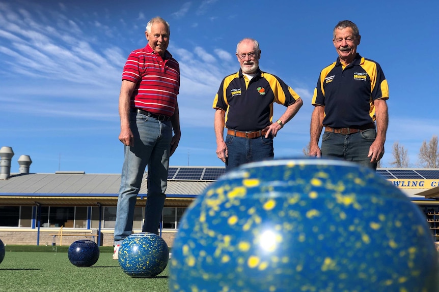 Three men stand on a green surface smiling with a big blue lawn bowl in front of them.