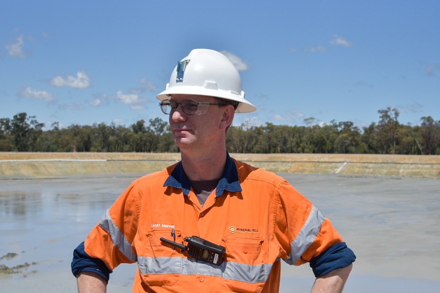 A man in high vis with a white helmet and walkie talkie stands in front of a tailings dam