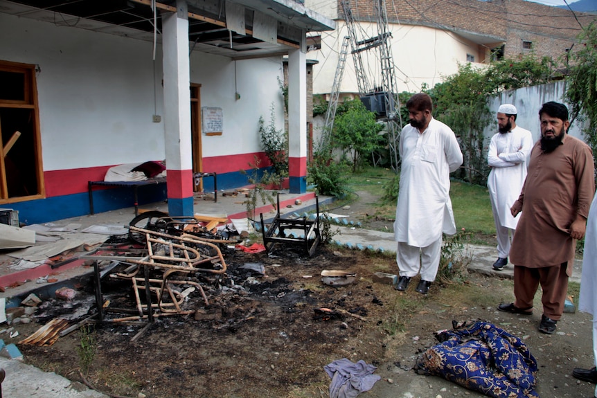 Three men in plain Pakistani garb look at piles of burnt furniture outside a building.