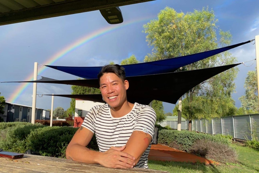 Mr Tran-Van sitting at a park bench with a rainbow in the background. 