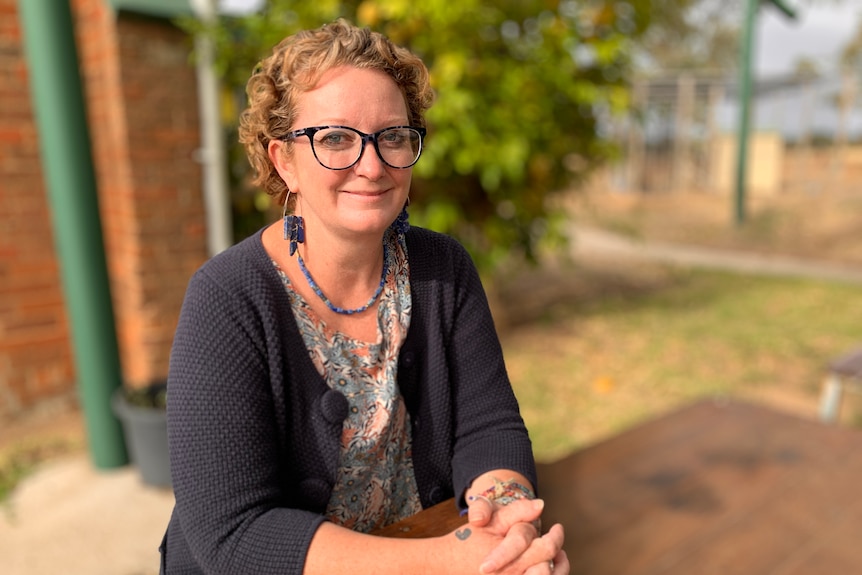 Woman wearing a printed dress and cardigan and glasses, sitting at a schoolyard table.