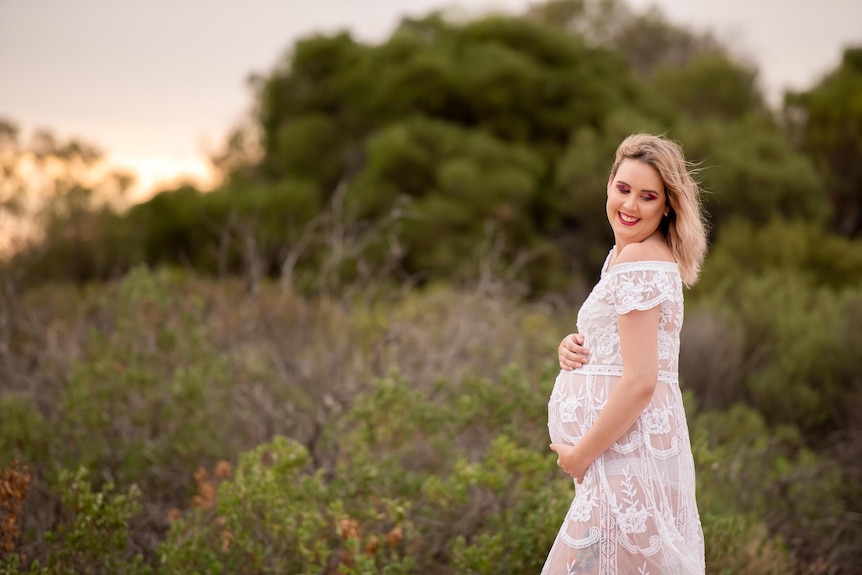 A women in a white dress holds her baby bump happily.