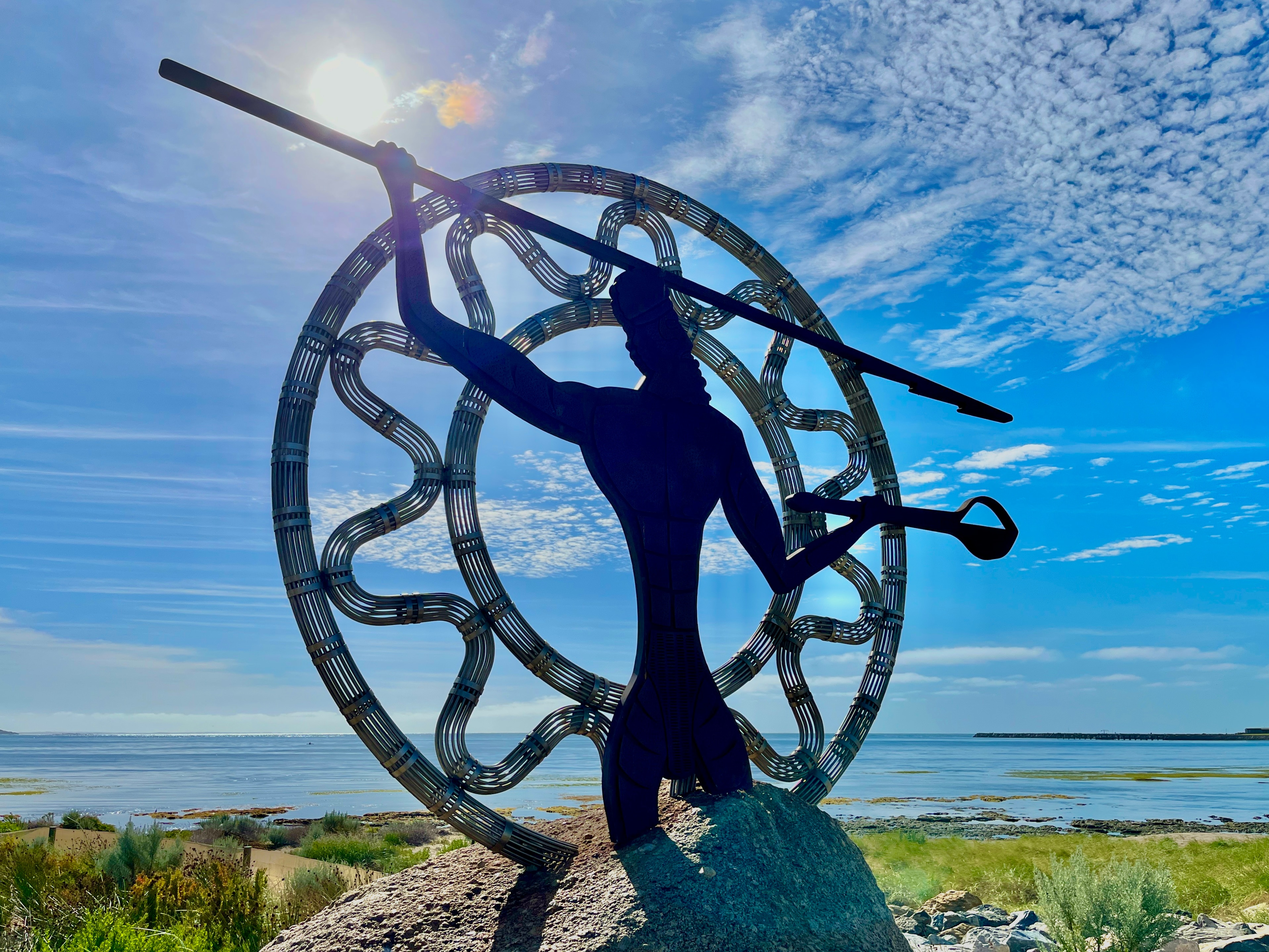 A metal statue of a warrior with raised spear and club with the background of the sun embedded into a large granite boulder