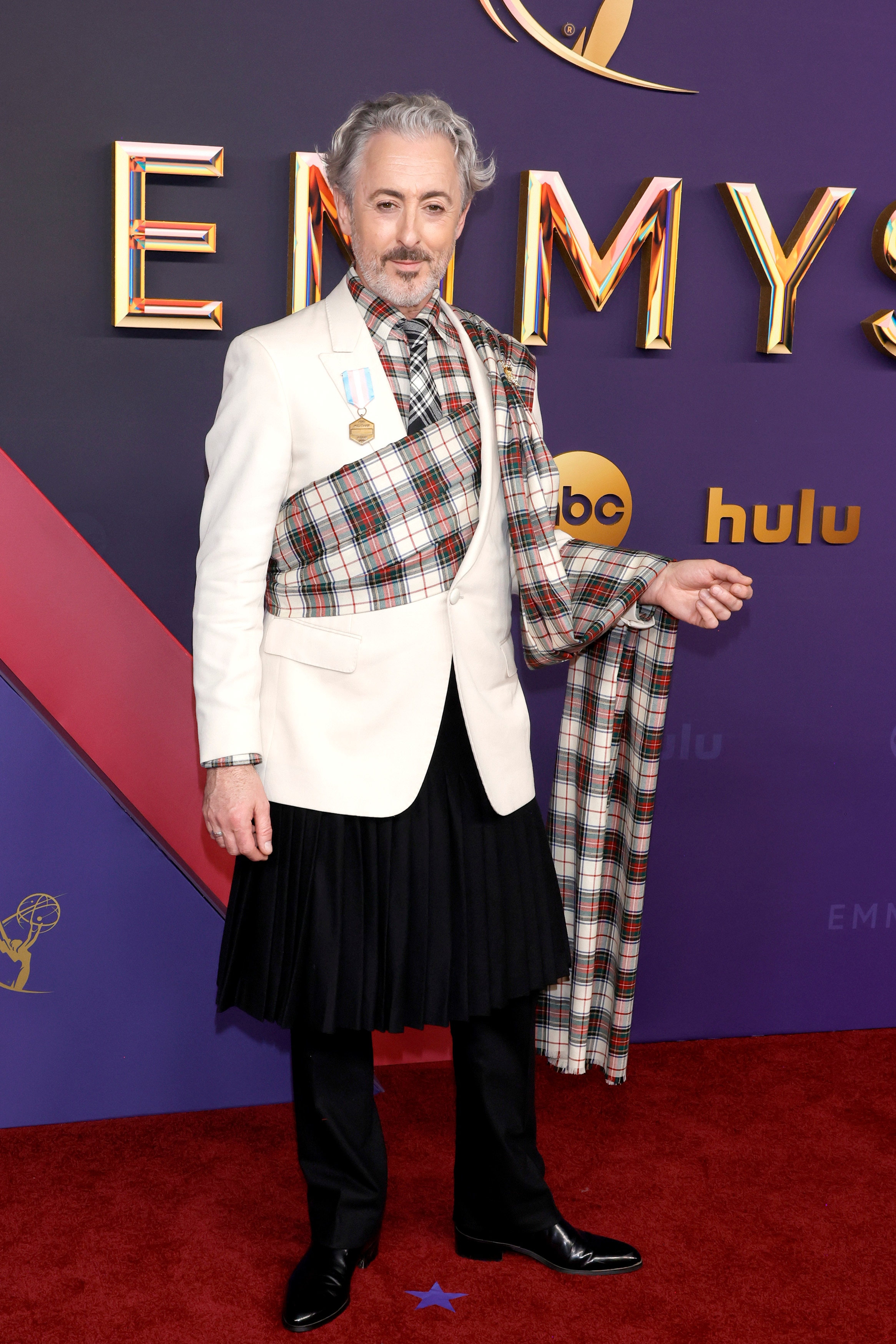 Alan Cumming wearing black trousers with a white dinner jacket and a red and white tartan scarf draped around his torso