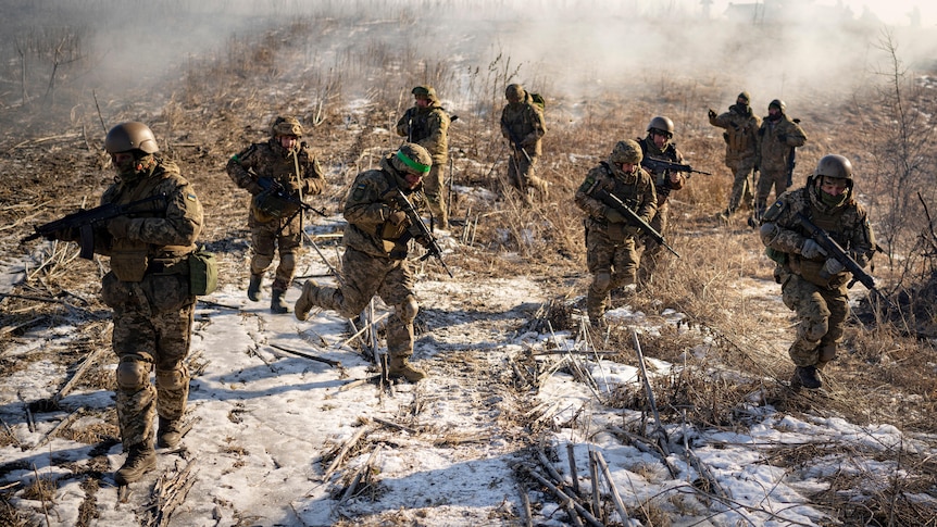Ten soldiers move across a barren snowy field with rifles raised.