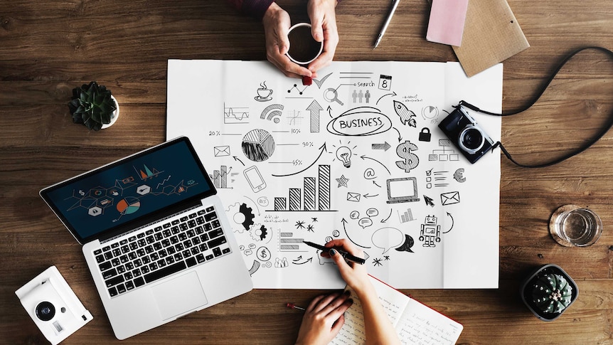 Overhead shot of a laptop, business plan, camera and hands around a coffee mug.